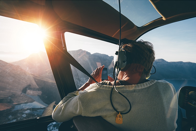 Rear view of female tourist on helicopter tour taking pictures while flying over mountains on a sunny day.